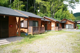 ウッドデッキがある黒色の三角屋根の木造平屋の短期滞在施設が4軒、手前から奥へ1列に並んでいる写真