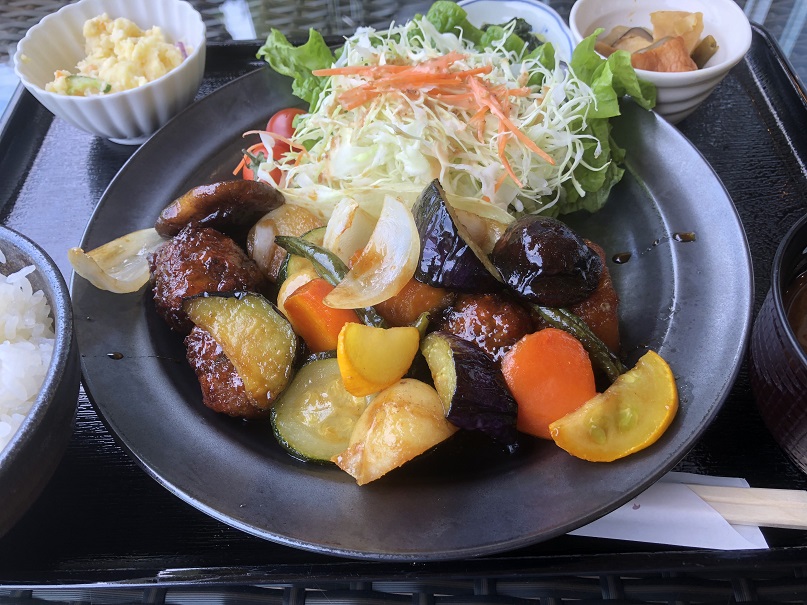 黒い皿に香酢で和えられた茄子、玉ねぎ、チョウザメだんご、人参と、千切りされたキャベツが添えられ、その四隅に小鉢に入ったポテトサラダなどの小鉢がある「チョウザメだんごの香酢定食」の写真