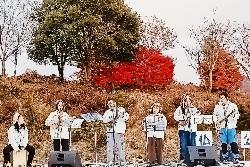 紅葉した草木をバックに5人のウクレレ奏者と1人の打楽器奏者が演奏している写真