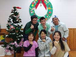 後ろに3人の男性、前に3人の女性がポーズを取っている。左側にクリスマスツリー、後ろにクリスマスのリース飾りがある写真。