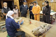 机の周りに6名の参加者が集まり、机に座っている男性が粘土を広げながら説明している様子を男性の後方から写した写真