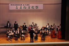 「とよね文化祭」と書かれた横断幕が掲げられたステージ上で、黒の衣装を着たオーケストラの方々が楽器を演奏している写真