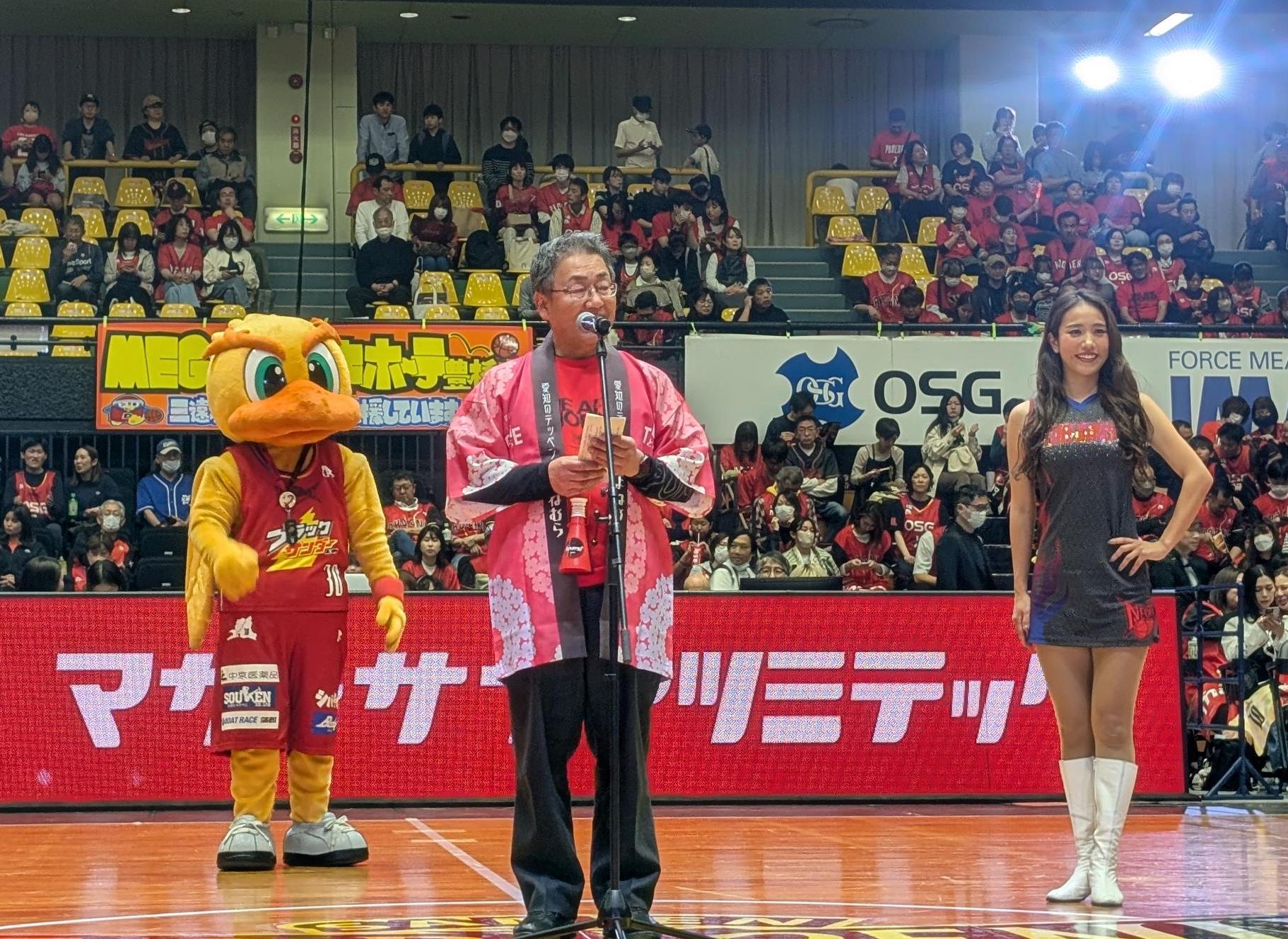 挨拶する村長の後ろに、チアガールとマスコットキャラクターがいる写真