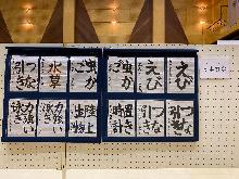 パネルに12枚の習字の作品が並んでいる。つな引き、水草、虫かご、えび、力強い泳ぎ、陸上生物、置き時計