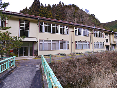 小さな橋の先にアイボリーを基調とした2階建校舎の坂宇場小学校の写真