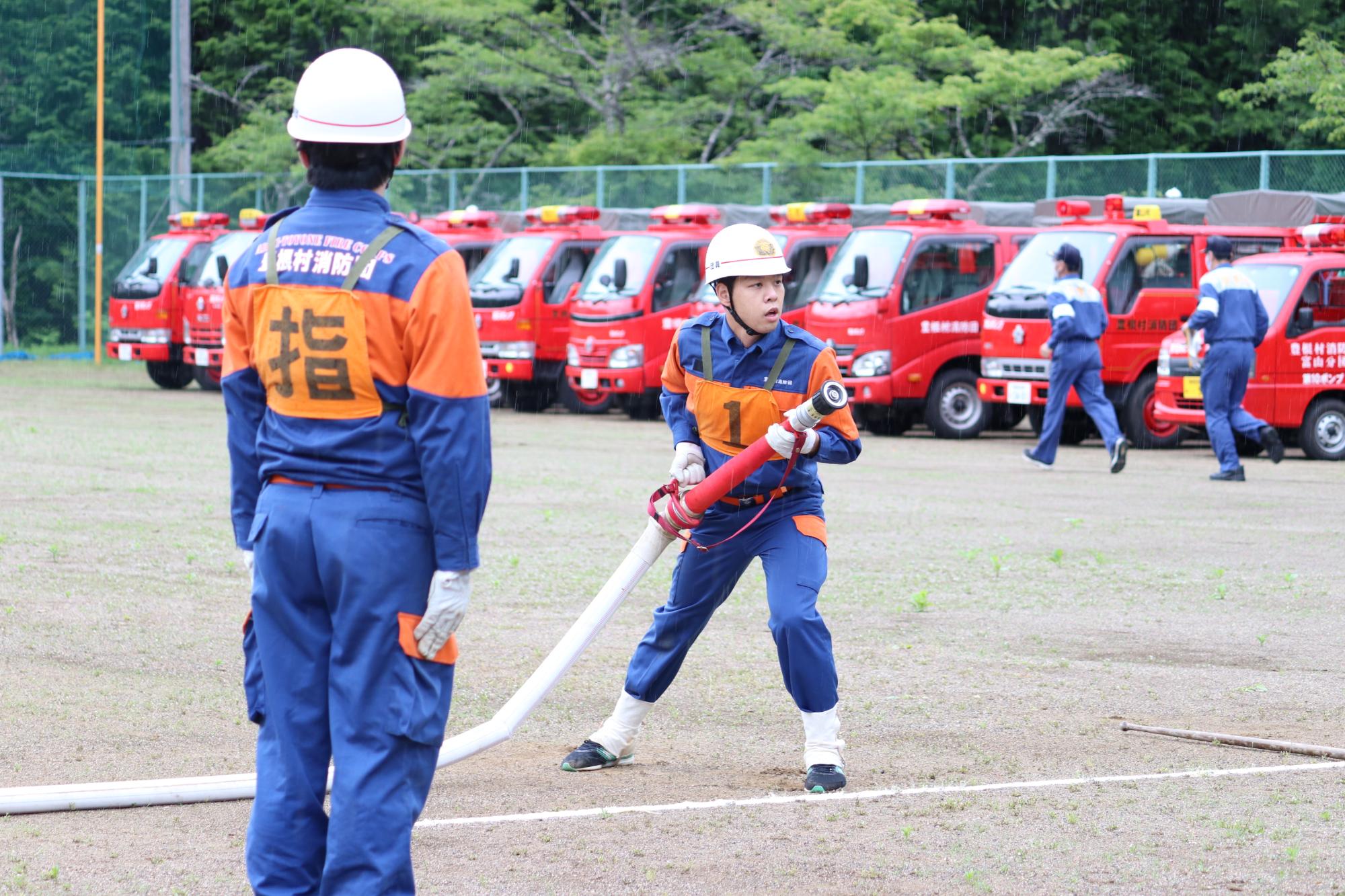 1名の消防団員がホースを持ち、手前のに指のゼッケンをつけた消防団員が指示を出しているの様子の写真