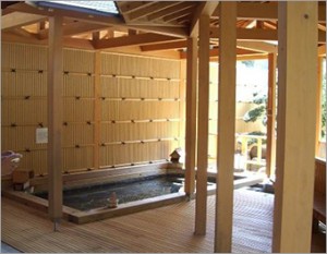 竹で出来た屋根付きの壁がある浴槽と右手には木があり、外からの光が差し込んでいる露天風呂の写真