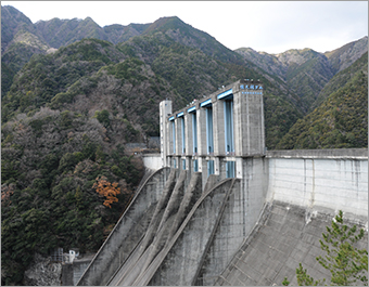 広がる山脈を背に、灰色と水色の急斜面の堤体が写っている「佐久間ダム」の写真