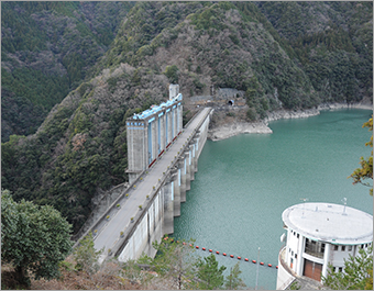 右側にエメラルドグリーンのダム湖があり、中央に建つ佐久間ダムを斜め上空から撮影した写真