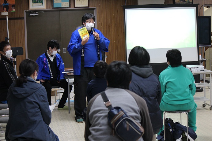 前方にスクリーンが設置された会場で青い半纏を着た男性が立って話をし、丸い椅子に座って話を聞いている参加者の方々の写真