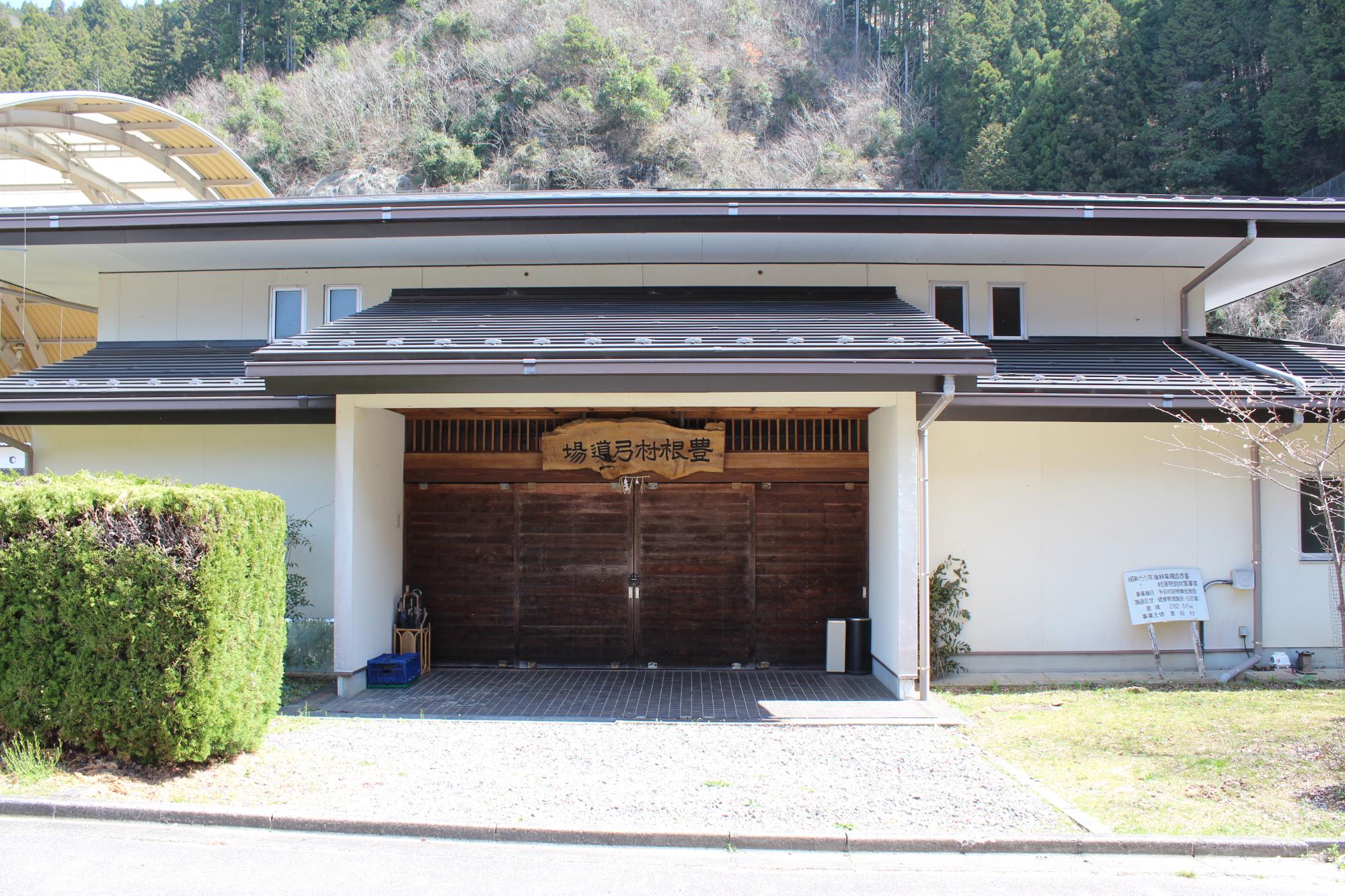 白を基調とし黒い屋根の建物で、「豊根村弓道場」と書かれた木の看板が設置されている豊根村弓道場の入り口の写真